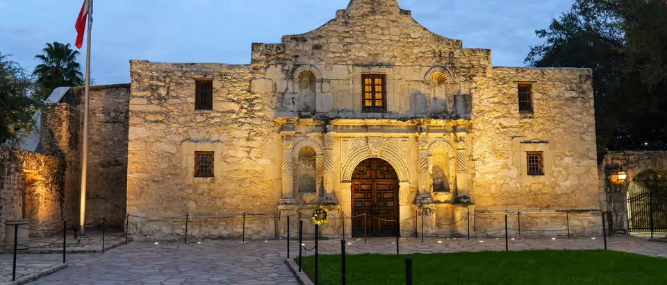 The Alamo's limestone church front lit at dusk under a blue sky, with a Texas flag on the pole at the left